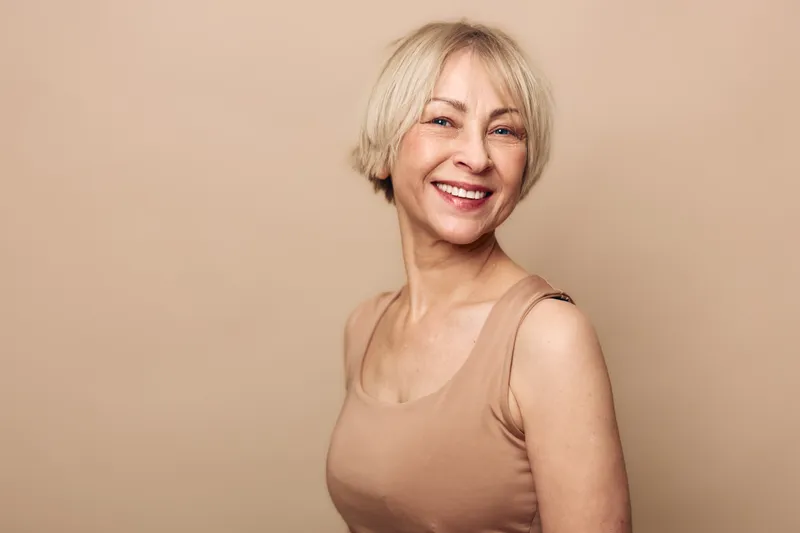 Smiling mature woman with short blonde hair wearing beige tank top poses against neutral background. Concept of confidence, aging gracefully, happiness, and natural beauty in lifestyle.