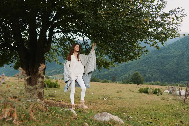 A woman in a light poncho stands beside a large tree in a rural field, posing with one arm raised. Calm lifestyle scene in nature, with soft light and open space.