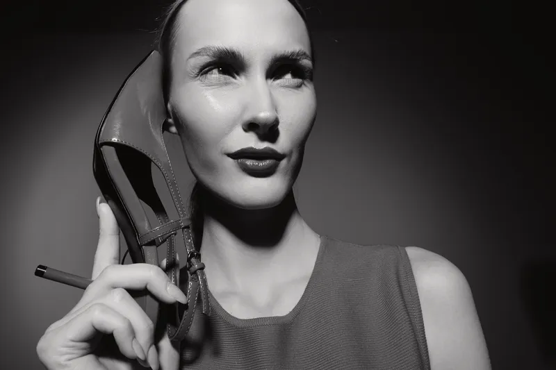 Portrait of caucasian woman with light skin holding high heel shoe near face. Black and white studio shot with dramatic lighting.