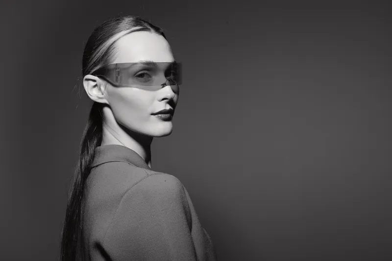 Young caucasian woman with pale skin and long hair wears modern transparent glasses in black and white portrait with dark background.