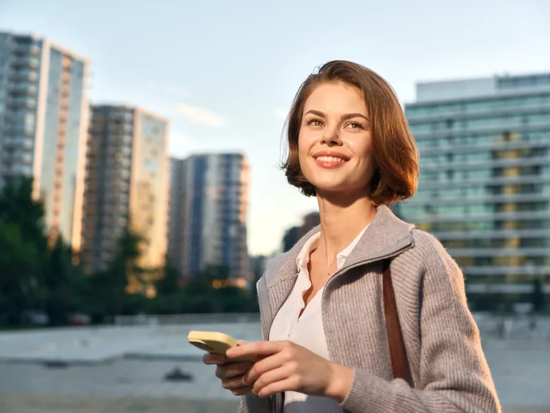 A cheerful professional woman stands outdoors in a modern city setting, holding a smartphone. She smiles brightly, appearing confident and engaged with work.