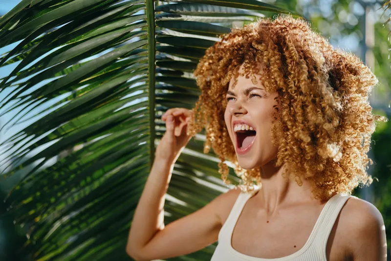 joyful woman with curly hair laughing outdoors near tropical palm leaves in natural sunlight, summer vibe, casual white tank top, energetic mood