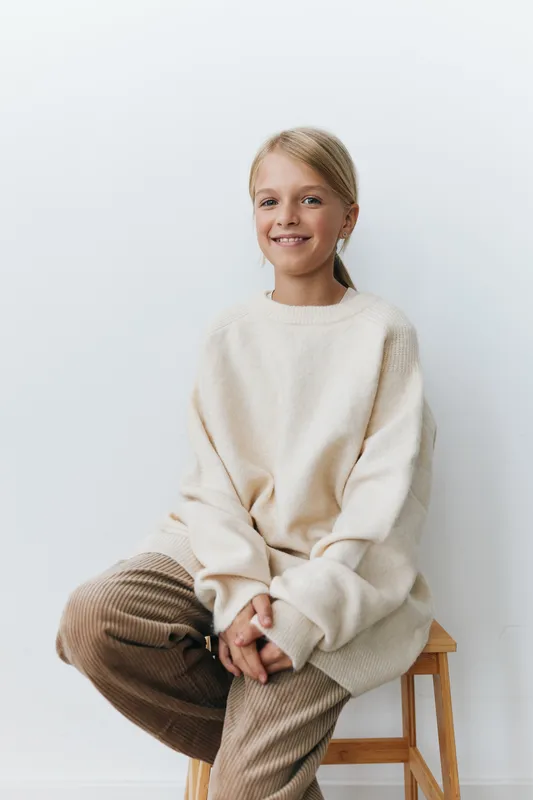 Young girl sits on a wooden stool wearing an oversized cream sweater and corduroy pants, smiling softly against a clean light background, conveying warmth and casual comfort.