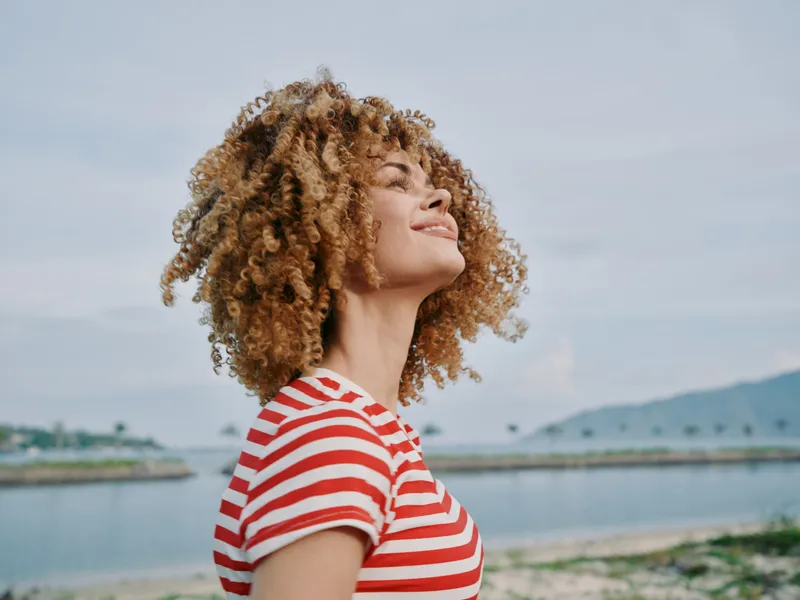 Joyful portrait of a young woman with curly hair wearing a red and white striped shirt, standing outdoors by a calm coastal backdrop with sunlight on her smile.