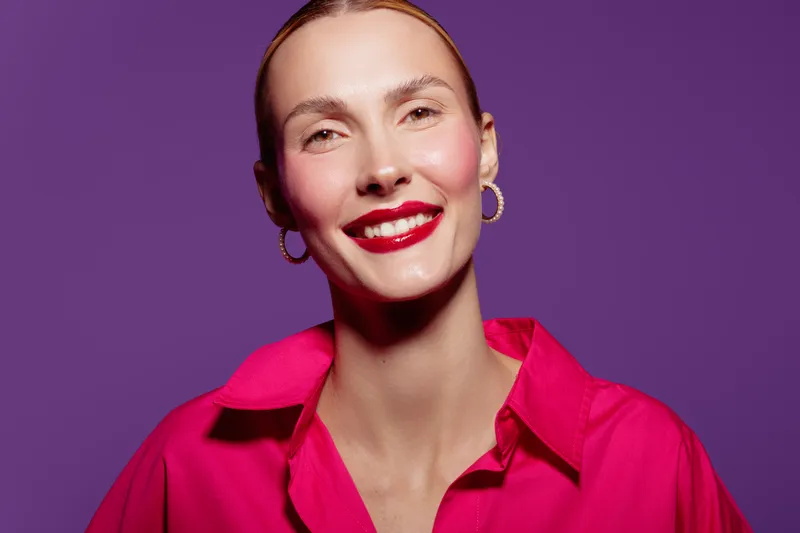 Smiling woman with red lipstick and gold hoop earrings wears bright pink shirt against purple background. Studio portrait with soft lighting highlights natural skin and confident expression.
