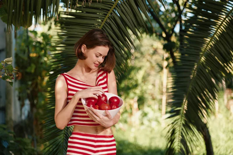 A cheerful woman wearing a red striped outfit holds a bowl of apples in a sunlit garden, smiling softly as she admires fresh produce, radiating health, vitality, and a simple wholesome lifestyle.