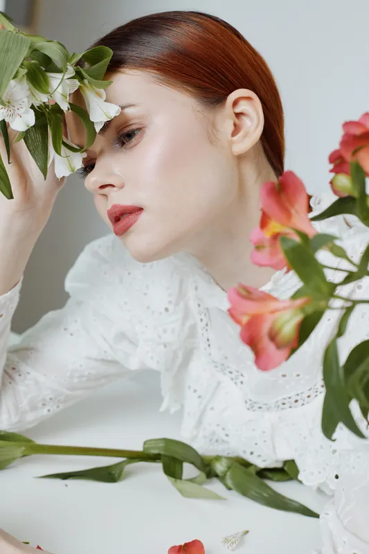 Delicate portrait of a woman with red hair, wearing a lace blouse, surrounded by colorful flowers, evoking beauty and femininity in soft lighting