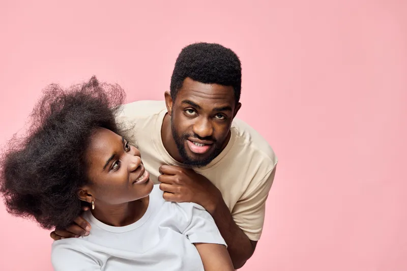 Happy young African couple posing together with playful expressions against a soft pink background, showcasing natural hair and stylish casual wear
