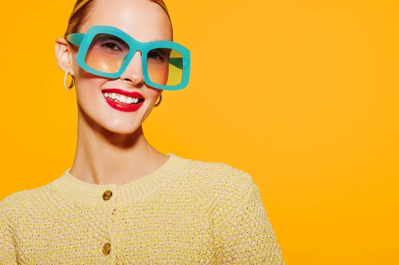 Smiling woman with bright red lipstick wears large turquoise sunglasses and yellow knitted sweater on vibrant orange background. Concept of fashion, style, and summer accessories.