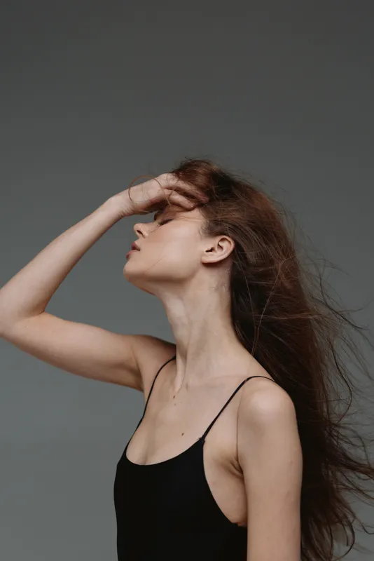 Profile of a young woman with long hair posing elegantly against a gray background in a black sleeveless top.
