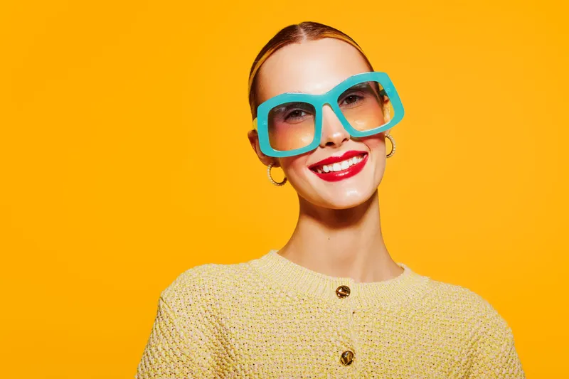Smiling woman with bright red lipstick wearing large blue sunglasses and yellow sweater on vibrant orange background. Concept of fashion, summer style, and confident mood.