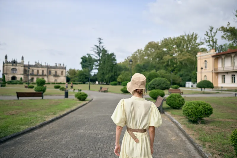 Woman in a vintage dress and hat walks along a gravel path in an expansive park, her back turned, creating a peaceful timeless scene near a grand estate.