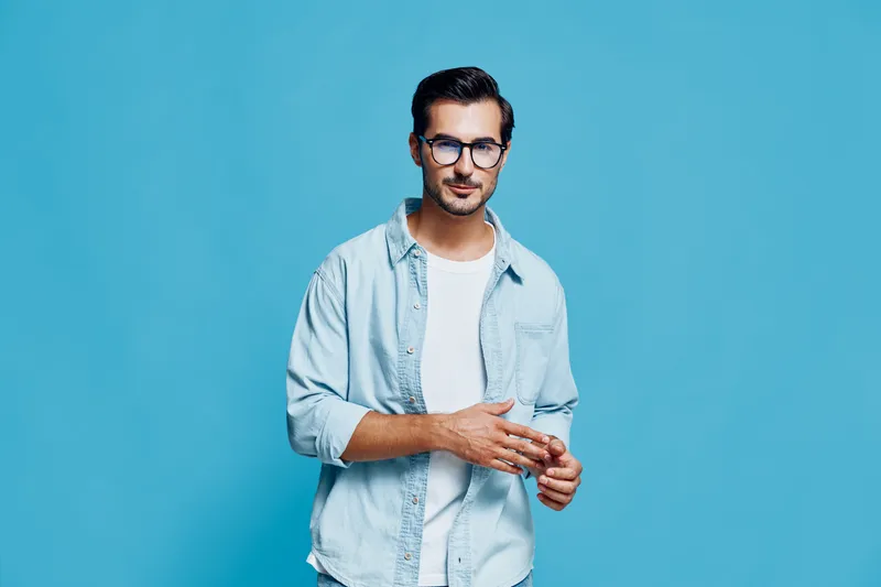 Stylish young man in casual light blue shirt and glasses stands confidently against a vibrant blue background, looking at the camera with a thoughtful expression.