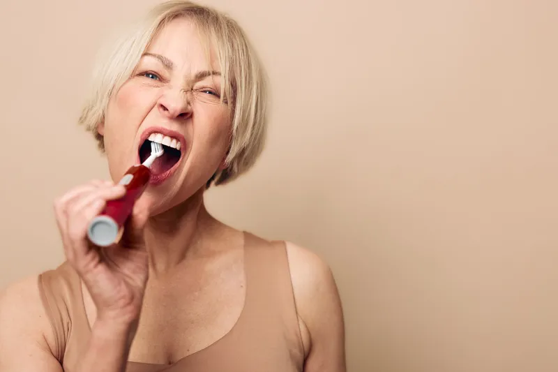 Mature woman with short blonde hair aggressively brushes teeth with electric toothbrush on beige background. Concept for dental hygiene, oral care, and personal routine.
