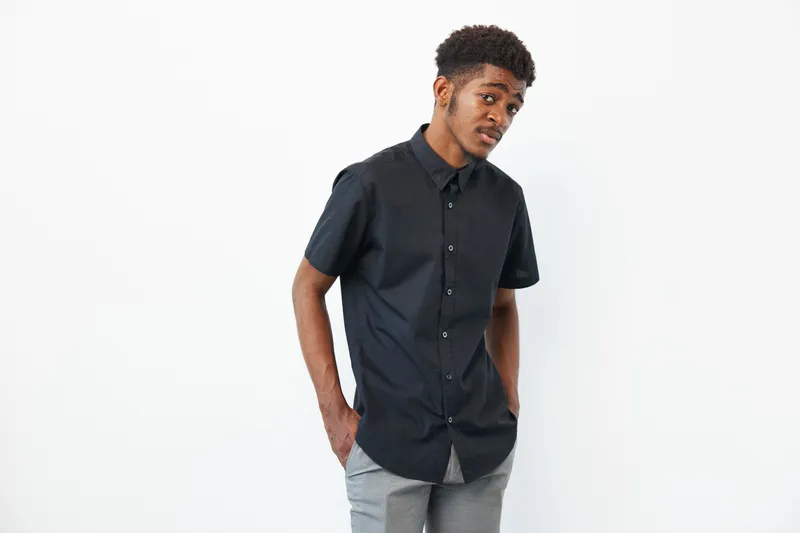 A young African American man poses in a stylish black shirt, showcasing confidence and modern fashion. The clean background emphasizes his trendy look and youthful energy.