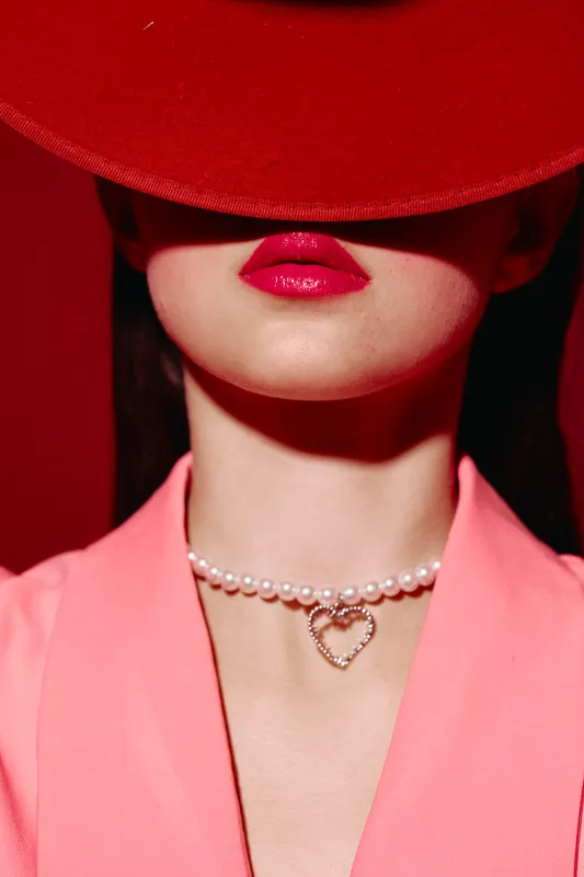 Close-up of woman wearing a red hat and pink blazer, featuring glossred lips and pearl necklace with a heart pendant. Creative beauty fashion portrait with dramatic vivid colors.