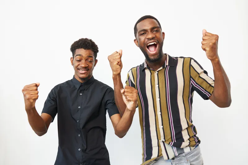 Two African American men celebrating with joy, showing excitement and happiness in their expressions, wearing fashionable casual outfits and posing together.