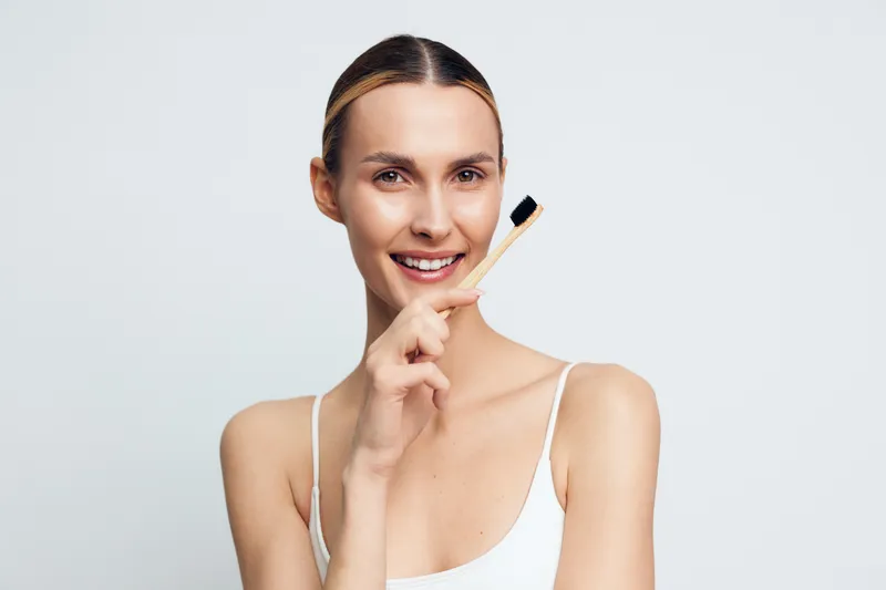 Smiling woman holding bamboo toothbrush near mouth on white background. Concept of dental care, oral hygiene, natural lifestyle, and eco friendly habits. Soft lighting highlights fresh mood.