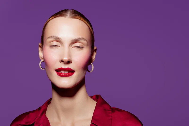 Portrait of a woman with red lipstick and hoop earrings wearing a red blouse on purple background. Concept of beauty, fashion, makeup, and confident female model with closed eyes.