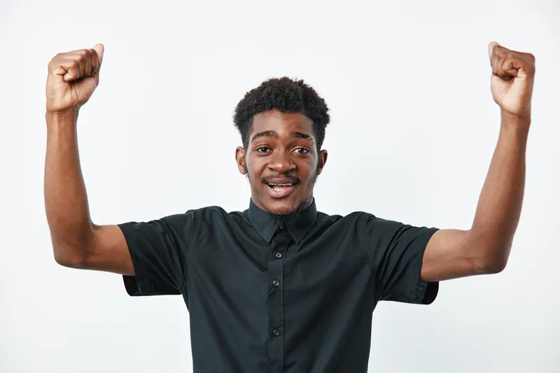 Young black man with dark skin wearing black shirt raising fists in celebration or victory isolated on white background. African ethnicity, happy expression and casual style.