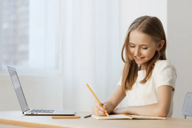 Cheerful young girl writing in a notebook at a bright desk with a laptop, capturing the essence of creativity and focus in a minimalistic workspace