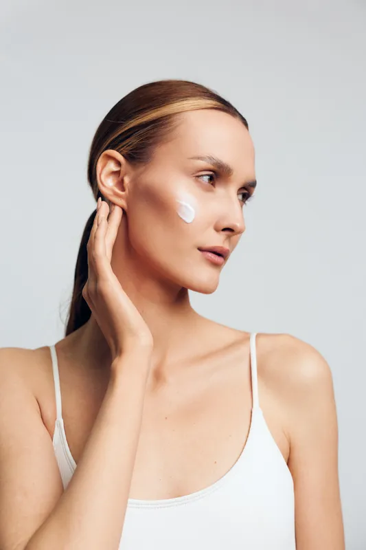 Woman skincare applying cream face close up profile portrait, young woman spreads white cream on cheek, soft studio lighting on clean white background, concept of self care and beauty