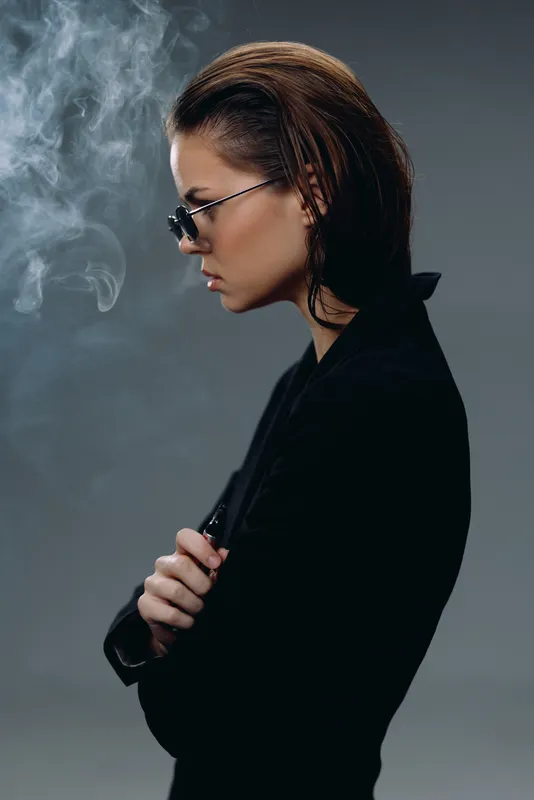 Profile of a thoughtful woman wearing glasses surrounded by smoke in a dark, moody atmosphere.
