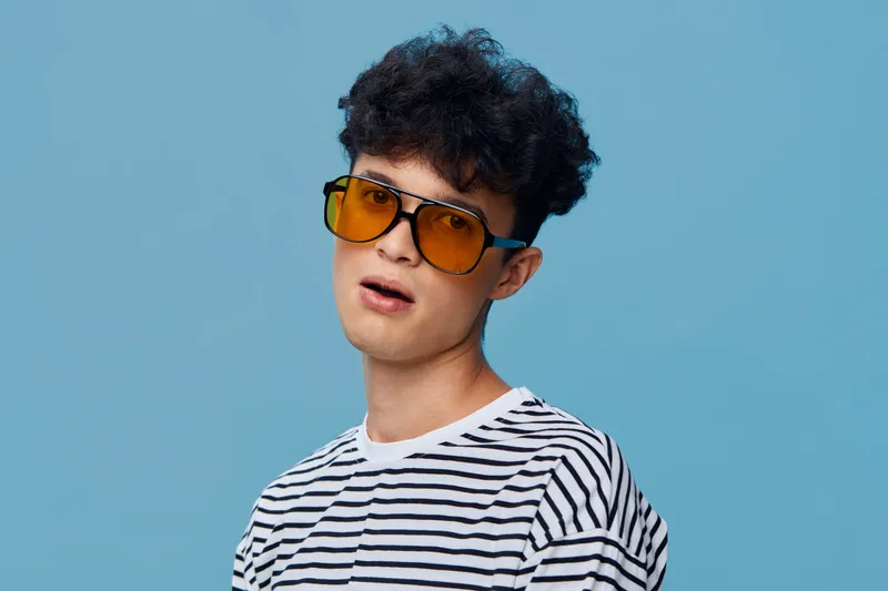 young man with sunglasses wearing striped shirt posing against blue background, casual style, dark curly hair, youthful expression, minimalism, studio lighting