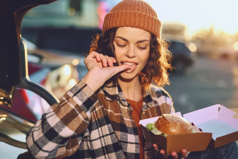 Woman eating burger from a box by a car trunk outdoor, beanie and coat, candid lifestyle shot capturing authenticity, golden hour glow, mindful living and emotional storytelling.