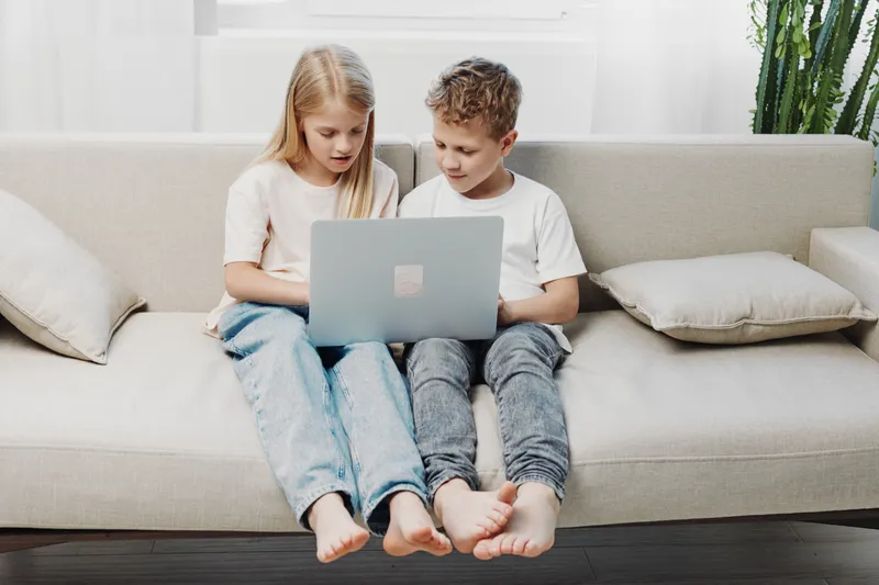 Two barefoot children sitting on a beige couch using a laptop together, focused on the screen in a bright living room with neutral decor and cushions.