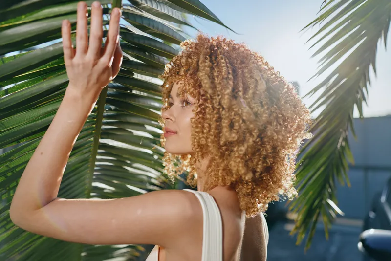 Curly woman standing near palm leaves in sunny outdoor setting, wearing sleeveless top, natural light highlighting her hair and serene expression.