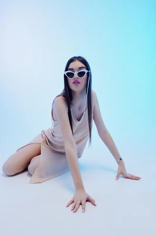 Stylish woman with long braided hair wearing white sunglasses and a beige dress poses on the floor against a light blue background. Concept for beauty, fashion, and style.