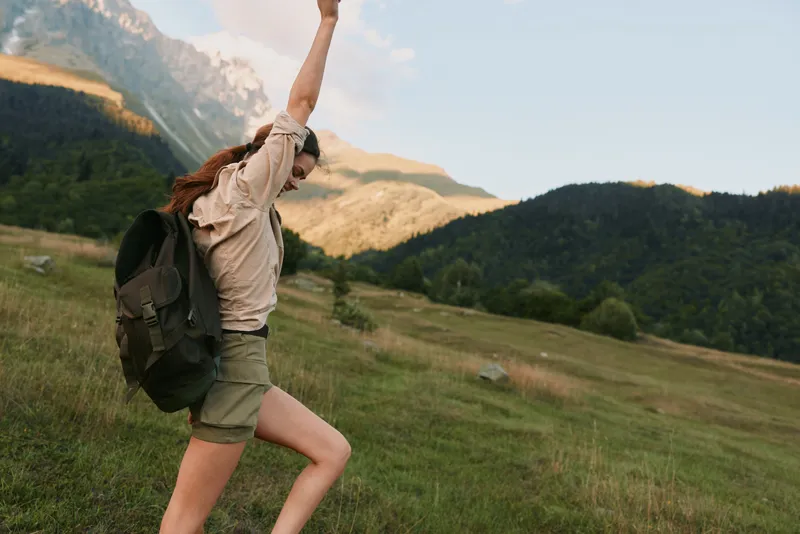 Woman hiker with backpack raises arms in a sunlit meadow, mountains in the background, portraying freedom, energy, and outdoor lifestyle during a hiking day.