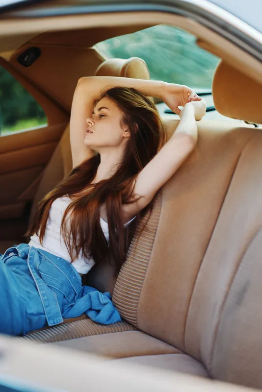 Young woman relaxing in a car with long hair, wearing a white top and denim jeans, conveying a sense of freedom and serenity in a natural setting.