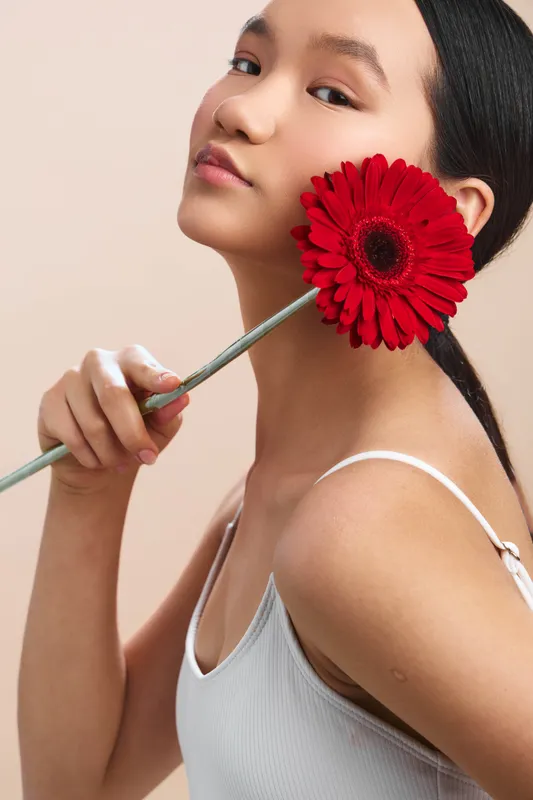 Asian woman holding a vibrant red flower near her face in a soft studio setting. She gazes gently toward the camera, conveying calm elegance and contemporary beauty.