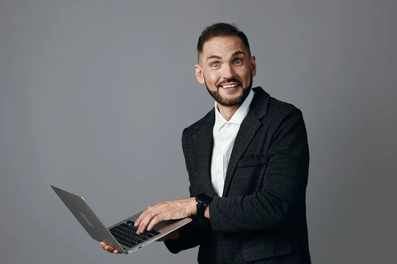 A professional man in a tailored blazer smiles while using a laptop, conveying confidence, energy, and focus against a solid colored background.