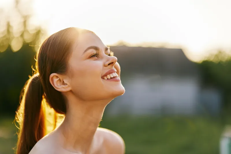 Young woman with long hair smiling joyfully outdoors during sunset, radiating happiness and warmth in a lush green garden