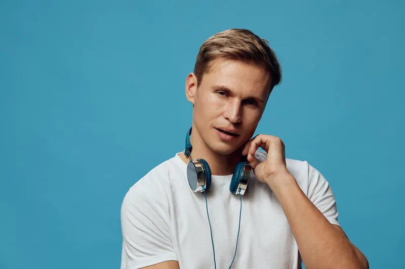 Smiling man with headphones around neck in casual white t-shirt posing against blue background. Studio portrait of cheerful young male with friendly expression and modern style. People lifestyle conce