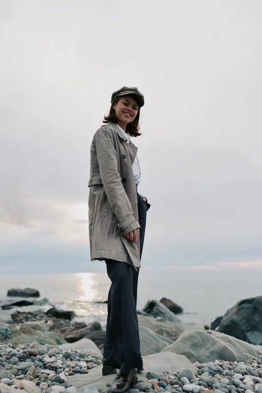 Smiling woman in casual coat and cap stands on rocky beach near calm sea during cloudy sunset. Concept of leisure, travel, autumn fashion, and outdoor lifestyle.