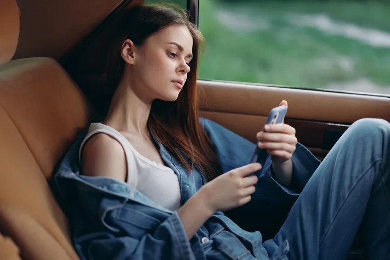 Young woman in casual denim outfit relaxing in car, using smartphone, expressing calmness and comfort, surrounded by natural greenery.