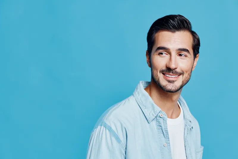 Smiling man in denim shirt looking over shoulder with cheerful expression against a solid blue background in a studio setting. Personality and style concept.