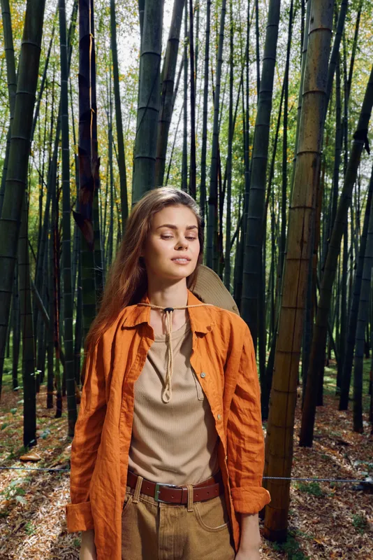 Woman bamboo forest portrait with eyes closed and hiking backpack, orange jacket in sunlight, nature travel scene showing serene outdoor meditation, peaceful eco tourism and relaxed lifestyle.