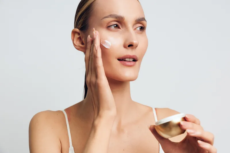 Woman applying face cream with fingers and holding a jar for skincare self care beauty Close up portrait on a light studio background Soft studio lighting