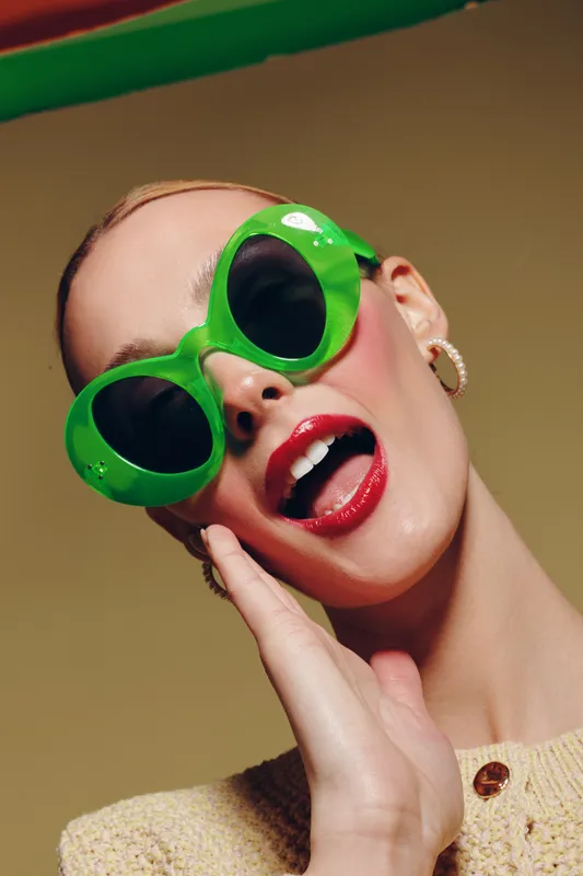 Close-up of a fashionable woman wearing large green sunglasses and red lipstick, posing with an open mouth and hand near face. Concept of style, beauty, and summer accessories.