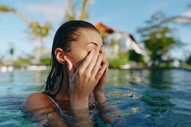 Young woman in a pool rinses her face with water, hands covering features, bright outdoor setting with tropical vibe, lifestyle moment of refreshment and self care during vacation