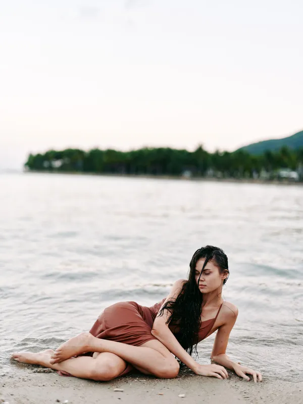Female figure lounging on a sandy shore beside calm water, wearing a brown swimsuit, long dark hair draped over shoulder, relaxed pose, soft light, tranquil coastal mood.