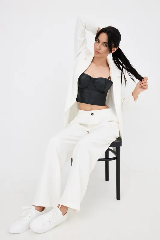 Young woman with long dark hair wearing white suit and black crop top sitting on chair, stylish casual outfit, confident pose on white background, modern fashion look.