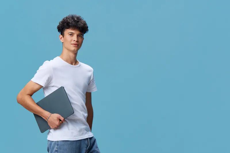 young man holding laptop in white t-shirt standing against blue background, casual style and confident expression in modern technology concept