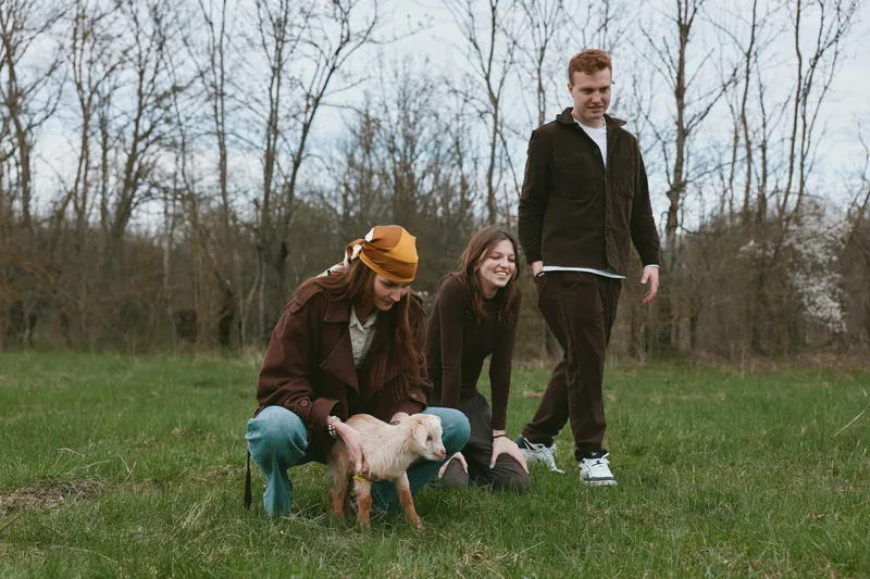 Three friends enjoy outdoor time with a playful goat kid in a grassy field, capturing a casual lifestyle moment with gentle animal interaction and warm friendship.