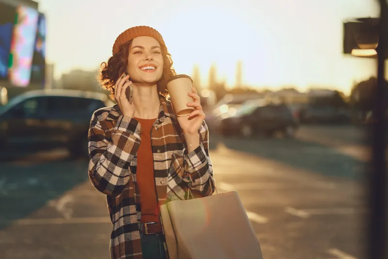 Woman with coffee and shopping bag smiling in urban sunlight, candid lifestyle portrait at golden hour glow capturing authenticity, mindful living and emotional storytelling.